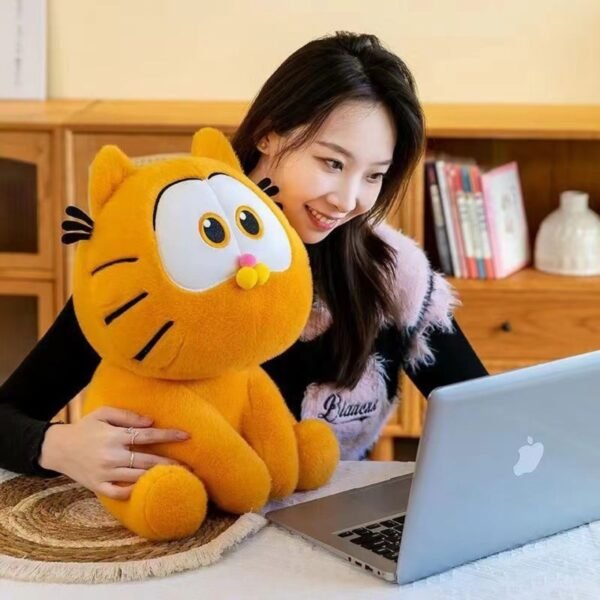 Smiling woman with long hair holding a large orange Garfield plush toy while looking at a silver laptop on a table.